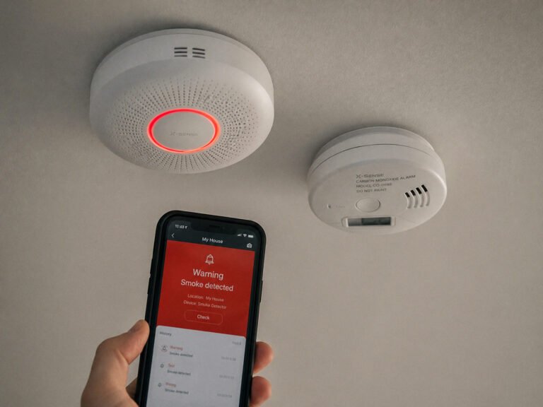 Smart smoke and carbon monoxide detectors mounted on a home ceiling with a phone alert on screen